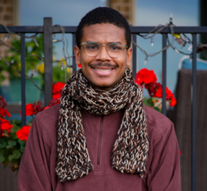 Photo of Aqshems Nichols smiling, wearing glasses, a brown woven scarf, and maroon pullover